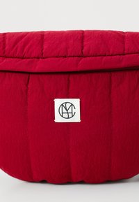 Borsa da stoccaggio imbottita rossa con texture trapuntata. Presenta un'etichetta bianca con un logo nero, corpo rotondo e un coperchio pieghevole.