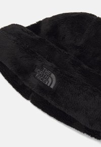 Sort fleece-lue i svart med en myk tekstur, rund form og et logo-merke med "The North Face" i grått på siden.