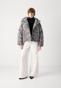 Giacca padronale con un motivo zigzag bianco e nero, texture trapuntata, colletto oversize e zip frontale, abbinata a pantaloni bianchi a gamba larga.