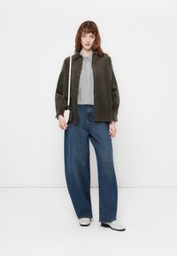 Olivově zelená oversized košile, černobílý pruhovaný top pod ní, široké modré džíny a bílé boty s malou crossbody taškou.