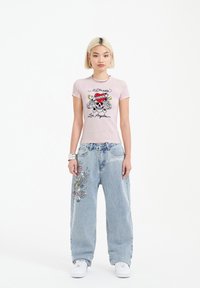Light pink fitted t-shirt with skull graphic and text; paired with loose blue denim jeans featuring multicolored embroidered designs. White sneakers.