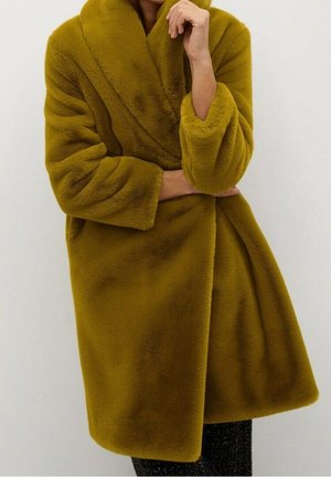 Donna che indossa un cappotto di pelliccia sintetica color senape con collo a scialle, tenendo il cappotto chiuso con una mano vicino al viso.
