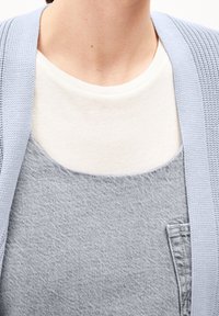 Vaaleansininen neulemainen neuletakki valkoisen paidan päällä, yhdistettynä harmaisiin haalareihin, joissa on rintatasku ja teksturoitu kangas.