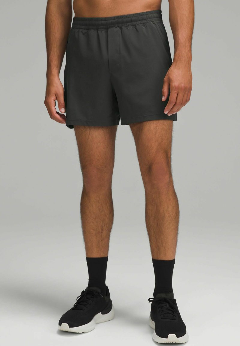 Shorts de sport noirs avec une ceinture élastique, fabriqués en tissu léger. Le modèle porte des baskets noires et des chaussettes.