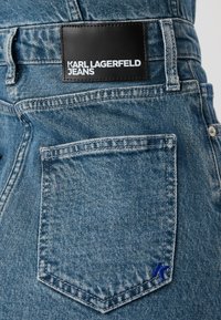 Išblukę mėlyni džinsai, turintys juodą odos aplikaciją su užrašu "KARL LAGERFELD JEANS" ir mažu mėlynu logotipu ant užpakalinio kišenės.