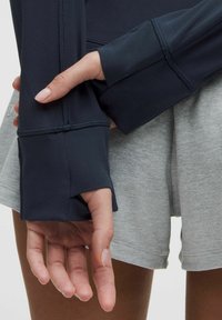 Jūras spēku zila garām piedurknēm sporta krekls ar pirkstu caurumiem, apvienots ar gaiši pelēkiem šortiem. Iezīmē plakanšuvju šuves un mīkstu, elastīgu audumu.