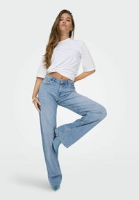 Jeans a gamba larga di colore blu chiaro con orlo grezzo, abbinati a una maglietta bianca a maniche corte, modella in posa con una gamba alzata su uno sfondo neutro.