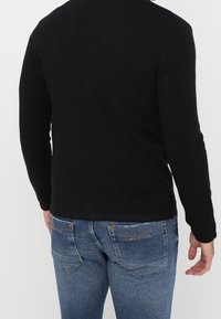 Musta, pitkähihainen neule, valmistettu teksturoidusta kankaasta, yhdistetty sinisiin farkkuihin, joissa on takataskut ja vaaleat sauma-yksityiskohdat.