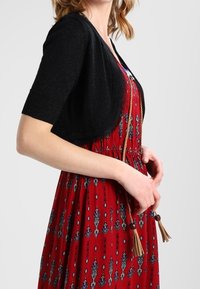Czarny bolerko z krótkim rękawem na czerwonej sukience z niebiesko-białymi wzorami; posiada wiązanie w talii z chwostami. Materiał o fakturze.