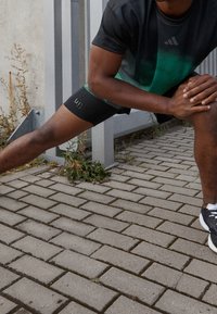Svart och grön gradient träningströja, åtsittande svarta shorts med logo, sträckpose, liggande på en asfalterad yta med grönska.