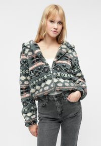 Šedá mikina s nápletom z umelej kožušiny, zdobená geometrickými vzormi v krémovej a broskyňovej farbe. Zkrátený strih s kapucňou a elastickými manžetami.