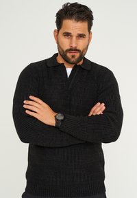 Schwarzer gestrickter Pullover mit Kragen, der eine gerippte Textur und ein tailliertes Design aufweist. Das Modell trägt eine Uhr am linken Handgelenk.