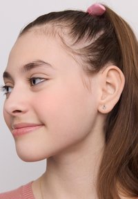 Złota kolczyk wkręcany z małym, przezroczystym akcentem kamienia szlachetnego. Modelka ma długie brązowe włosy i różowy sweter, co wyraźnie eksponuje kolczyk.