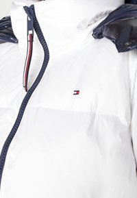Biała kurtka puchowa z wysokim kołnierzem, granatowym kapturem i granatowym suwakiem. Posiada małe logo na klatce piersiowej i paskowy akcent na suwaku.