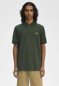 Polo verde con maniche corte, caratterizzato da un colletto con tre bottoni e un logo in oro a forma di alloro sul lato sinistro del petto.