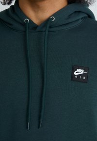 Sweatshirt verde escuro com cordão, apresentando um patch preto quadrado no peito com texto do logo em branco. Material em algodão, textura suave.