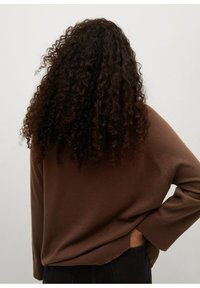 Osoba z długimi kręconymi ciemnymi włosami, ubrana w brązowy sweter, z jedną ręką na biodrze, widziana od tyłu na jednolitym jasnym tle.