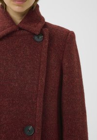 Cappotto di lana in un ricco colore bordeaux, presenta un motivo a spina di pesce testurizzato, un grande bottone nero e un colletto alto per maggiore calore.