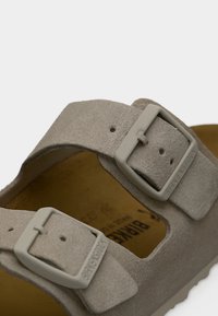 Κοντινό πλάνο μπεζ σουέντ σανδαλιού Birkenstock με δύο ρυθμιζόμενα λουριά και ματ αγκράφες πάνω σε ταμπά πέλμα.