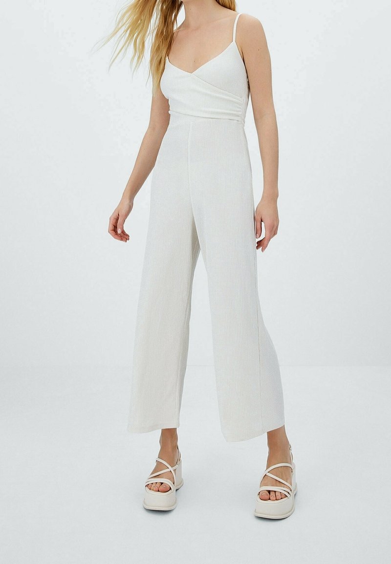 Femme portant une combinaison sans manches blanche avec des jambes larges et des sandales à plateforme crème, se tenant devant un fond clair uni.