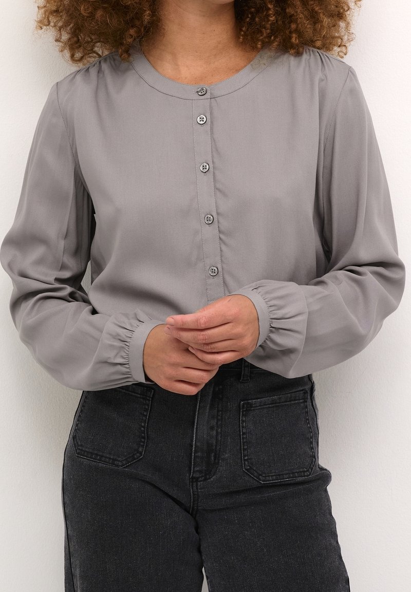 Femme portant un chemisier gris clair boutonné à manches longues et un jean noir taille haute avec poches avant, les mains jointes devant.
