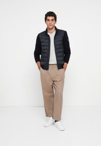 Jeune homme standing, portant une veste quiltée noire avec des manches en tricot, un pantalon large beige, des baskets blanches, les mains dans les poches, fond neutre.