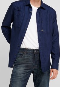 Marineblauwe button-up shirt met een gestructureerde afwerking, twee borstzakken en zwarte knopen, gecombineerd met donkere spijkerbroeken.