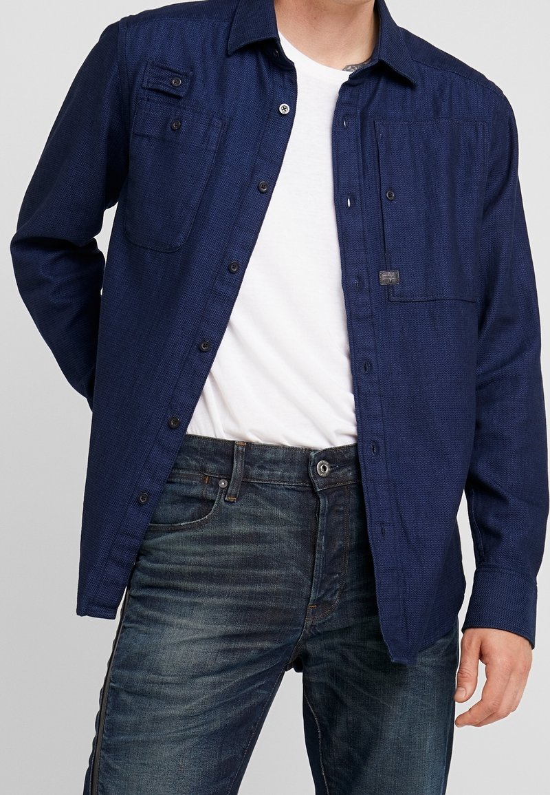 Marineblauwe button-up shirt met een gestructureerde afwerking, twee borstzakken en zwarte knopen, gecombineerd met donkere spijkerbroeken.