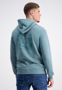 Mīksta kapuce viegli tējas krāsā, izgatavota no kokvilnas materiāla. Aizmugurē ir reljefa grafiska druka, ribota aproces un apakšmala.