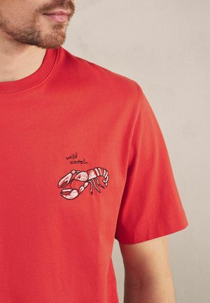 Man wearing red T-shirt with embroidered lobster and text "wild catch" on left chest, plain light background