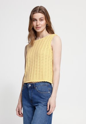 Junge Frau mit hellbraunen Haaren, die ein ärmelloses gelbes Strickoberteil und blaue Jeans trägt, steht vor einem schlichten hellen Hintergrund.