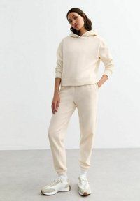 Sudadera de color crema y joggers a juego hechos de un material suave, con puños acanalados y un ajuste relajado, acompañados de zapatillas ligeras.