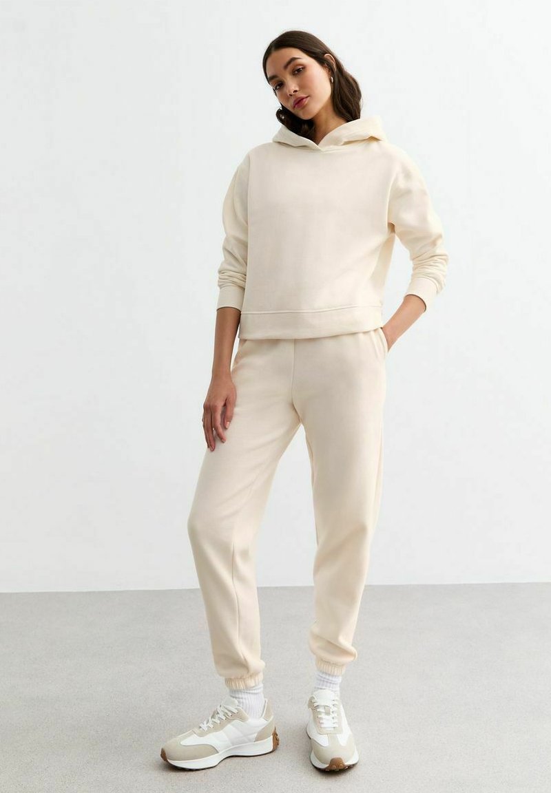 Sudadera de color crema y joggers a juego hechos de un material suave, con puños acanalados y un ajuste relajado, acompañados de zapatillas ligeras.