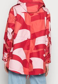 Set bagfra af en person iført en hættejakke med mønster i rød, pink og koral, med justerbar talje og knapper ved ærmerne, kombineret med lyseblå jeans.