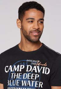 Svart t-shirt med djärv vit och blå text. Rund halsdesign med subtila ränder på axlarna. Slät, lättviktig tyg.