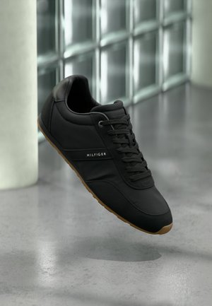 Fekete Tommy Hilfiger sportcipő barna talppal, megkötött fűzőkkel, szürke padló fölött lebegve, egy beltéri üvegfal közelében.