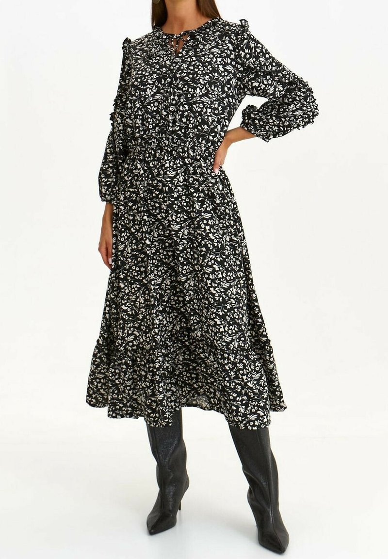 Femme portant une robe midi noire à imprimé floral blanc, manches longues froncées, et bottes noires à talons montantes jusqu'aux genoux, debout, main sur la hanche.