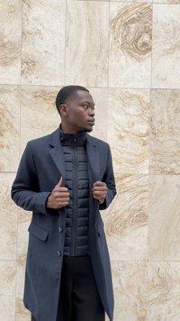 Man in dark coat and black quilted jacket standing against beige marble tile wall, looking to his left, holding coat lapels.
