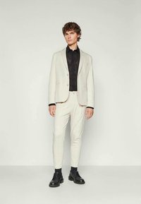 Młody mężczyzna stoi w jasnobeżowym garniturze, czarnej koszuli i czarnych butach, na jednolitym jasnym tle.