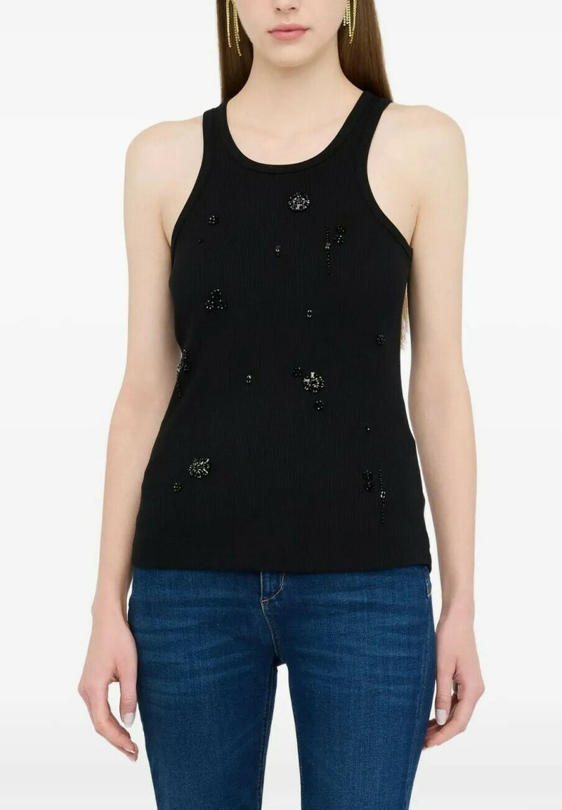 Donna con top nero senza maniche decorato con piccole perline nere e jeans blu, su sfondo bianco semplice.