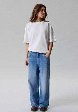 Kvinne iført en enkel hvit oversized T-skjorte, løse lyseblå jeans og mørkebrune sko, stående med en hånd i lommen.