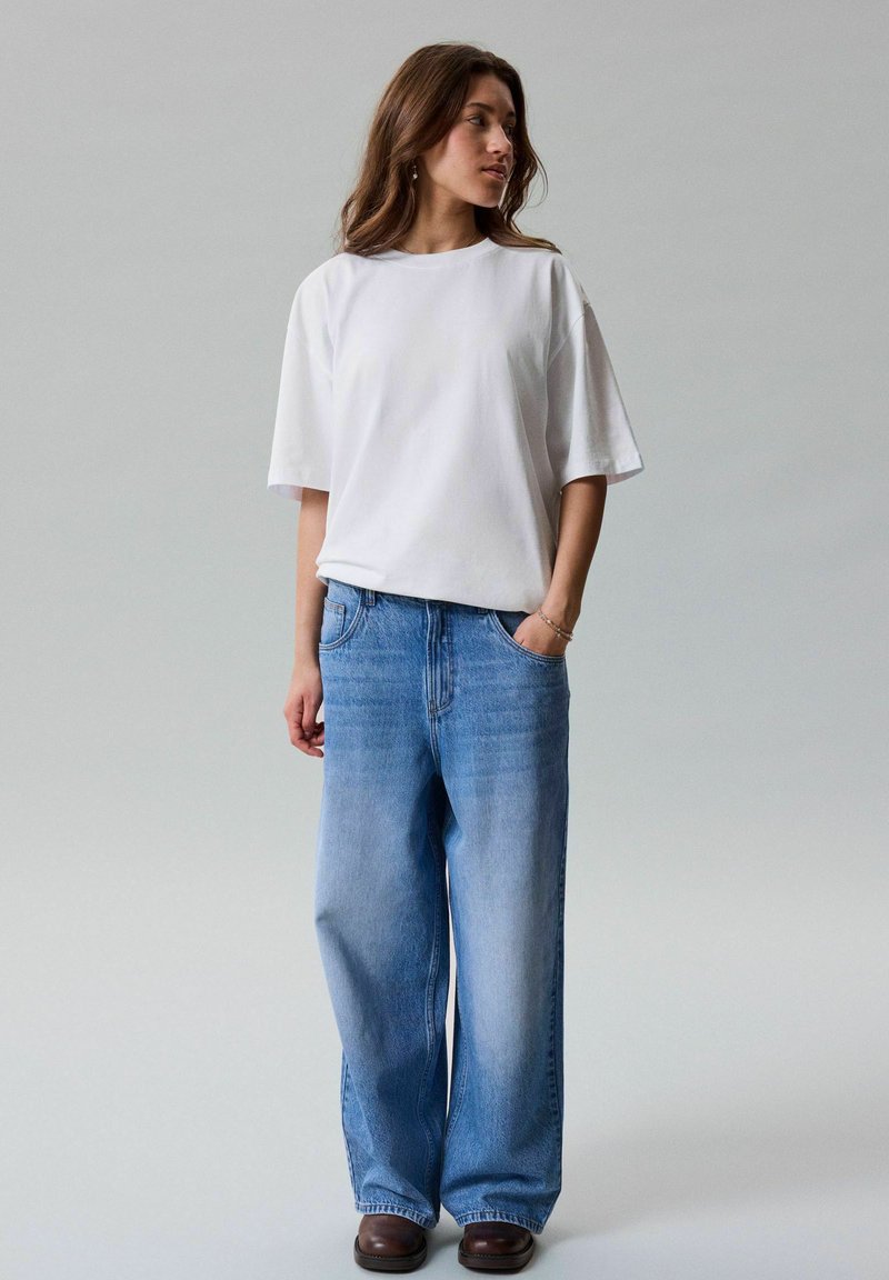 Γυναίκα που φοράει ένα απλό λευκό oversized T-shirt, φαρδιά ανοιχτό μπλε τζιν και καφέ σκούρα παπούτσια, στέκεται με το ένα χέρι στην τσέπη.