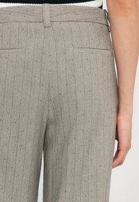 Harmaat herringbone-housut sisältävät hienovaraiset raidoitukset ja pieniä koristeellisia helmiä. Niissä on kaksi takataskua ja vyötäröllä vyö.