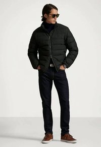 Άνδρας με μαύρο μπουφάν, σκοτεινά τζιν, καφέ αθλητικά παπούτσια, γυαλιά ηλίου και ρολόι, στέκεται με τα χέρια στις τσέπες σε απλή φόντο.