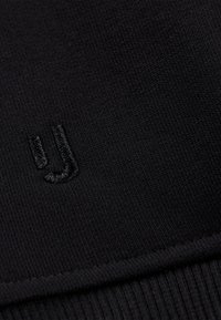 Tejido de punto negro con textura acanalada y un pequeño logo bordado en la esquina. Superficie suave con un brillo sutil.