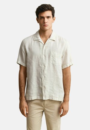 Mann steht vor einem schlichten Hintergrund, trägt ein hellbeiges Kurzarm-Hemd mit Brusttasche und beige Hosen.