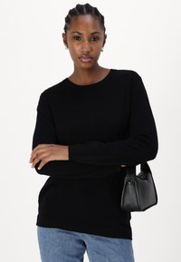 Black long-sleeve top with a round neckline, paired with light blue denim. A small black handbag is held under the arm.