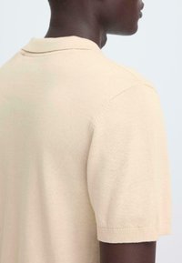 Polo beige en maille avec une texture lisse, manches courtes et col classique. Vue arrière mettant en avant des détails subtils de couture.