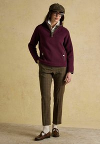 Sieviete, tērpta brūnās vairāk svītrainās biksēs, baltās zeķēs, brūnos loafers, bordo džemperī, baltā krekla, brūnā plakanā cepurē un brillēs, stāv pie bēša sienas.