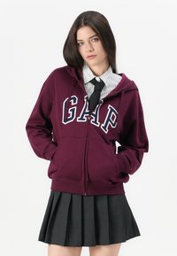 Young woman wearing a maroon GAP hoodie, white striped shirt with black tie, and a pleated dark gray skirt, standing with hands in pockets.